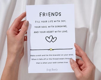 May include: A white card with the word "FRIENDS" in black lettering, held by hands. The card includes a message about joy, sunshine, and love, along with a black string bracelet with a gold star charm.