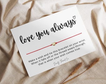 May include: A white card with the words "love you always" in black script, a red string bracelet with a gold heart charm, and text explaining the bracelet's purpose. The card is on a beige fabric.
