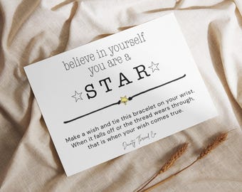 May include: A white card with the text "believe in yourself you are a STAR" and a black string bracelet with a gold star charm. The card also includes instructions and is set on a beige fabric background.