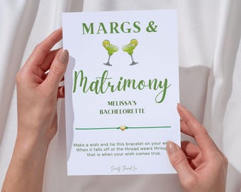 May include: A white card with green text and illustrations is held by hands. The card reads "MARGS & Matrimony MELISSA'S BACHELORETTE". Two margarita glasses are depicted above the word "Matrimony". A green string bracelet is attached.