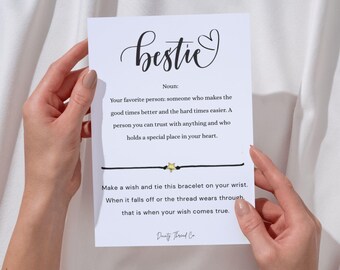 May include: A white card with the word "bestie" in cursive script, a definition, and instructions for a wish bracelet. The black bracelet has a gold star charm. The card is held by hands.