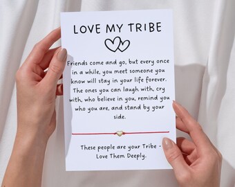 May include: A white card with the words "LOVE MY TRIBE" and a message about friendship. The card is held by hands and features a red string with a gold heart charm. The overall theme is friendship and love.