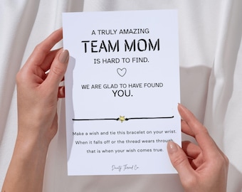May include: A white card with the text "A Truly Amazing Team Mom Is Hard to Find." A black string bracelet with a gold star charm is attached to the card. The card is held by two hands.