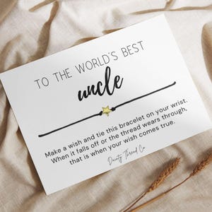 May include: A white card featuring the text TO THE WORLD'S BEST uncle with a black string bracelet and a small gold star charm. The card includes the printed message Make a wish and tie this bracelet on your wrist. When it falls off or the thread wears through, that is when your wish comes true. Dainty Thread Co is printed at the bottom.