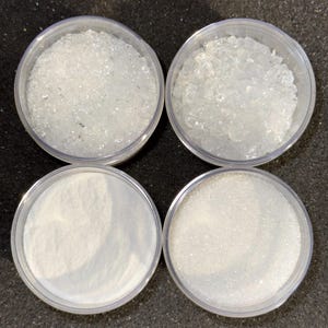 May include: Four clear plastic containers filled with different white substances. The top left container holds small, irregular crystals. The top right container has larger, more jagged crystals. The bottom left container contains a fine powder, and the bottom right has small, granular particles.