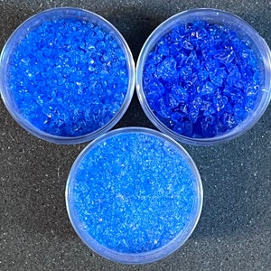 May include: Three clear glass containers filled with blue silica gel beads. The beads are small and irregular in shape.