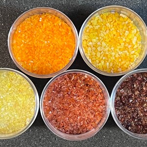 May include: Five small glass containers filled with different colored glass beads. The containers are arranged in a semi-circle on a dark gray surface. The beads are orange, yellow, light yellow, brown, and dark brown.