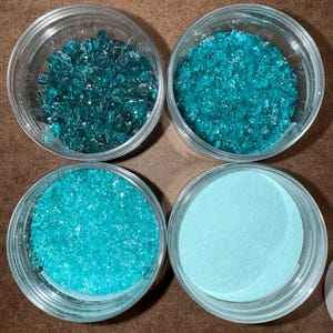 May include: Four clear plastic containers filled with different textures of teal-colored materials. The top left container holds chunky, irregular pieces. The top right has smaller, more uniform pieces. The bottom left has a granular texture, and the bottom right has a fine powder.