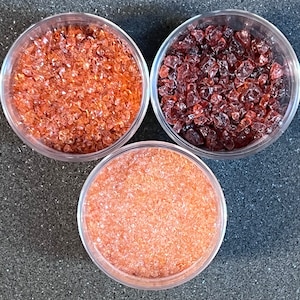 May include: Three clear, round containers filled with different colored, crushed glass. The top left container holds orange glass, the top right holds dark red glass, and the bottom container holds light orange glass. The containers are on a dark gray surface.