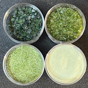 May include: Four clear glass jars filled with different shades of green glass beads. The beads are small and irregular in shape.