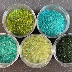 May include: Five small glass jars filled with different colored glass beads. The jars are arranged in a circle on a gray surface. The colors of the beads are green, teal, light green, and dark green.