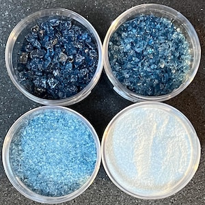 May include: Four clear glass jars filled with different shades of blue crushed glass. The jars are arranged in a square pattern on a dark surface.