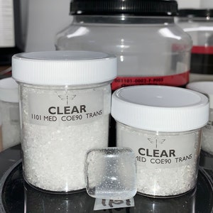 May include: Two clear glass jars filled with white crystals. The jars have white lids and labels that read "CLEAR 1101 MED COE90 TRANS".