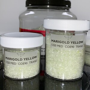 May include: Two small glass jars filled with translucent, light green glass frit. Both jars have white lids and labels that read "MARIGOLD YELLOW 1320 MED COE90 TRANS". A dragonfly logo is on each label.