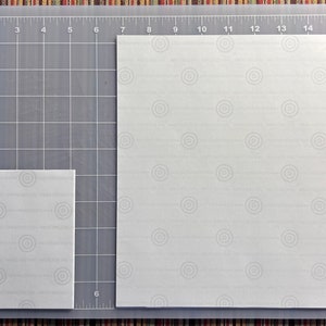 May include: Two white pieces of paper with a subtle pattern on a gray cutting mat with inch markings. The larger piece of paper is 12 inches by 16 inches and the smaller piece is 6 inches by 6 inches.