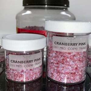 May include: Two clear glass jars filled with pink and white glass pieces. The jars have white lids and labels that read "Cranberry Pink 1311 Med COE90 Trans".