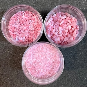 May include: Three clear glass jars filled with pink crushed rose quartz crystals. The jars are arranged in a triangle shape on a dark gray surface.