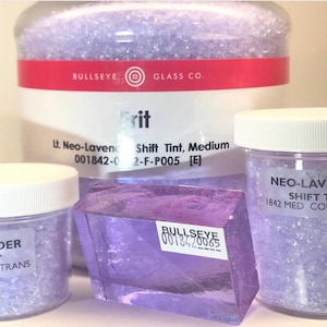 May include: Three jars of glass frit, a light purple color, labeled "Neo-Lavender Shift Tint" with the Bullseye Glass Co. logo. The frit is labeled with the product number 001842-0 and 1842. The frit is labeled as "Medium" and "COE90 Trans".