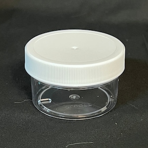 May include: A clear, small plastic jar with a white lid. The lid is round with a ribbed edge. The jar is empty and sits on a black surface. The jar is likely used for storing small items.