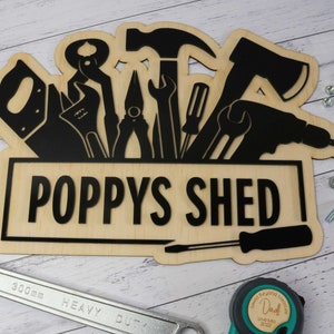 May include: Wooden sign featuring the text "POPPYS SHED" and a black silhouette of tools. The sign is light brown. A 300mm wrench, screws, and a tape measure are also visible.