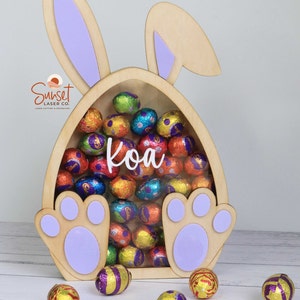 Pu&ograve; includere: Porta caramelle in legno a forma di uovo di Pasqua con orecchie e zampe di coniglio, pieno di ovetti di cioccolato avvolti in carta stagnola colorata. Il nome "Koa" &egrave; visualizzato sul davanti. Il logo Sunset Laser Co. &egrave; visibile.