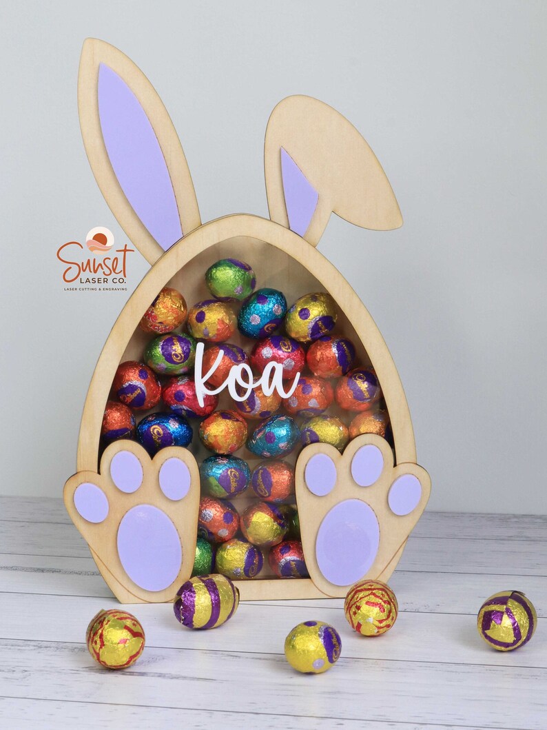 Pu&ograve; includere: Porta caramelle in legno a forma di uovo di Pasqua con orecchie e zampe di coniglio, pieno di ovetti di cioccolato avvolti in carta stagnola colorata. Il nome "Koa" &egrave; visualizzato sul davanti. Il logo Sunset Laser Co. &egrave; visibile.