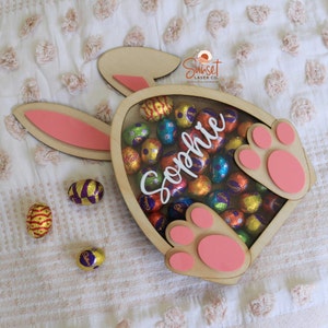 Pu&ograve; includere: Scatola di legno a forma di coniglio pasquale piena di ovetti di cioccolato avvolti in carta stagnola colorata. La scatola ha cuscinetti e orecchie rosa. Il nome "Sophie" &egrave; scritto in bianco. Diverse uova di cioccolato sono sparse.