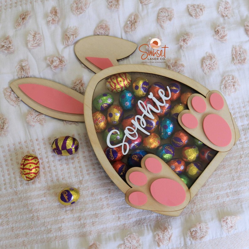 Pu&ograve; includere: Scatola di legno a forma di coniglio pasquale piena di ovetti di cioccolato avvolti in carta stagnola colorata. La scatola ha cuscinetti e orecchie rosa. Il nome "Sophie" &egrave; scritto in bianco. Diverse uova di cioccolato sono sparse.