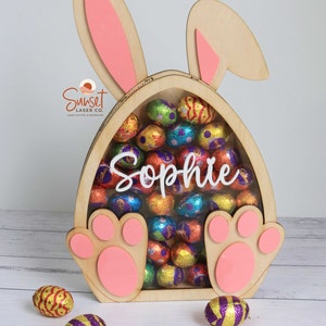 Pu&ograve; includere: Porta caramelle a forma di coniglietto pasquale in legno con dettagli rosa. Il coniglietto ha grandi orecchie e zampe a forma di zampa. Il supporto &egrave; pieno di uova di cioccolato colorate avvolte in carta stagnola. Il nome "Sophie" &egrave; visualizzato.