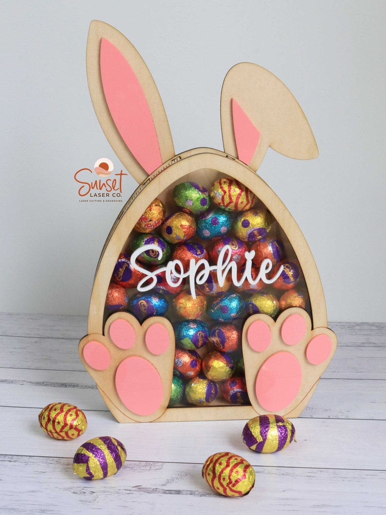 Pu&ograve; includere: Porta caramelle a forma di coniglietto pasquale in legno con dettagli rosa. Il coniglietto ha grandi orecchie e zampe a forma di zampa. Il supporto &egrave; pieno di uova di cioccolato colorate avvolte in carta stagnola. Il nome "Sophie" &egrave; visualizzato.