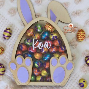 Pu&ograve; includere: Scatola di legno a forma di uovo di Pasqua con orecchie e zampe di coniglio, piena di uova di cioccolato colorate avvolte in carta stagnola. Il nome "Koa" &egrave; scritto in corsivo bianco. La scatola &egrave; circondata da altre uova avvolte in carta stagnola.