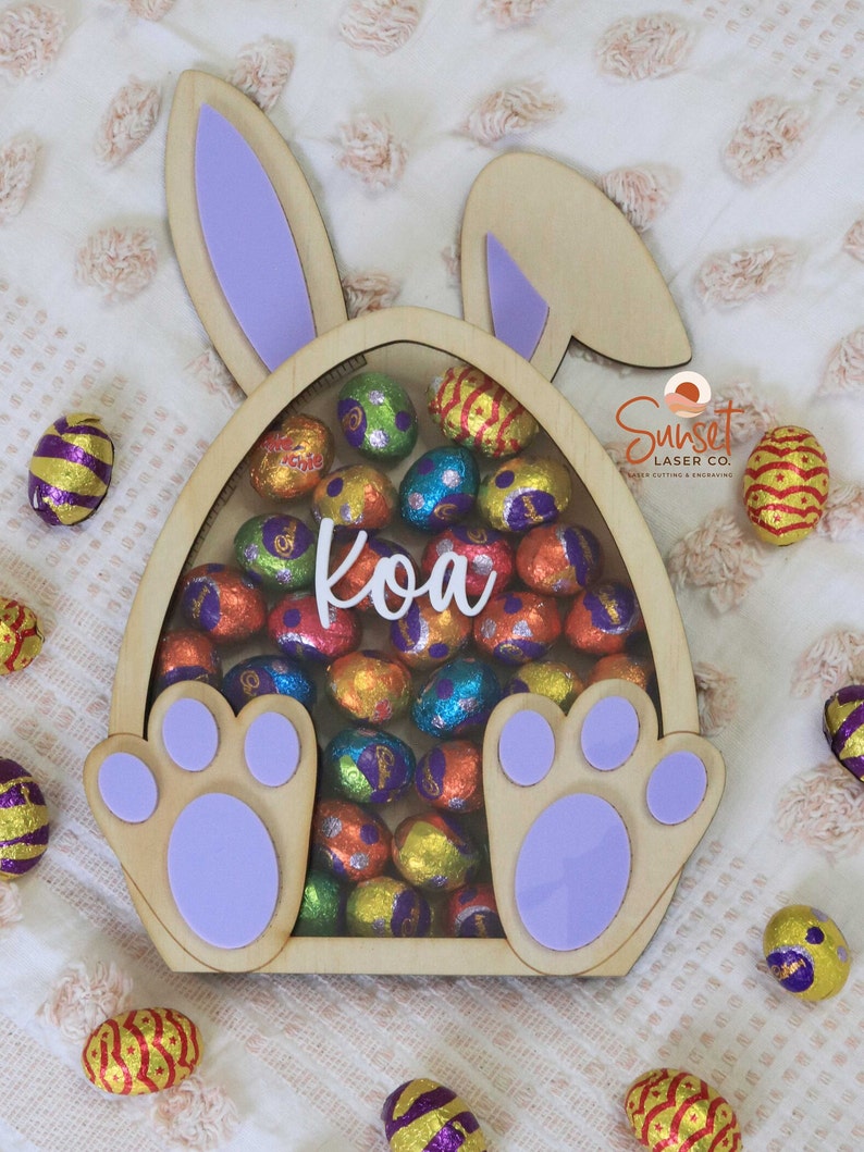 Pu&ograve; includere: Scatola di legno a forma di uovo di Pasqua con orecchie e zampe di coniglio, piena di uova di cioccolato colorate avvolte in carta stagnola. Il nome "Koa" &egrave; scritto in corsivo bianco. La scatola &egrave; circondata da altre uova avvolte in carta stagnola.