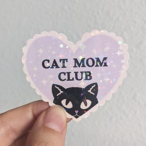 May include: A heart-shaped sticker with a scalloped edge featuring a light purple background with a holographic star pattern. The text CAT MOM CLUB is printed in black above a stylized black cat face with white accents.
