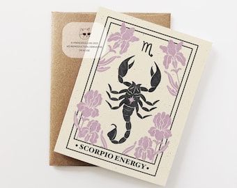 May include: A beige card featuring a black scorpion illustration with the text "SCORPIO ENERGY." The card is adorned with purple floral accents and a black border. A brown envelope is behind the card.
