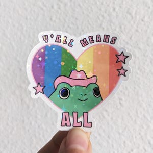 May include: A sticker featuring a green frog wearing a pink cowboy hat inside a heart-shaped design. The heart has a rainbow background and the words "ALL MEANS ALL" are printed around the heart.