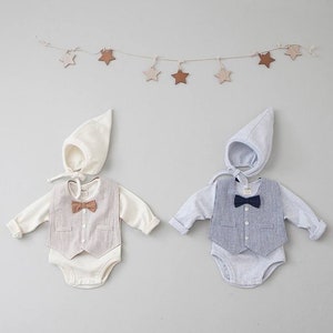May include: Two baby outfits with a vest and bow tie. One outfit is white with a brown bow tie and the other is gray with a navy blue bow tie. Both outfits have a matching white hat.