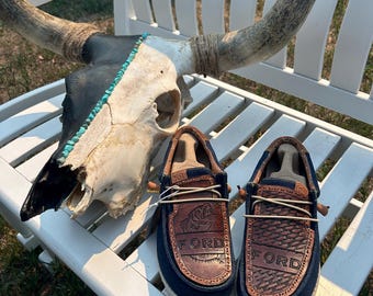 May include: A pair of blue canvas shoes with brown leather accents. The shoes have the word "FORD" embossed on the leather. The shoes are sitting on a white wooden bench with a cow skull with turquoise accents.