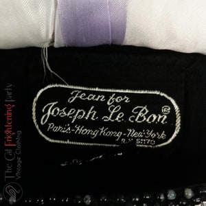 Pu&ograve; includere: Primo piano di un'etichetta di abbigliamento nera con testo ricamato bianco. L'etichetta recita "Jean for Joseph Le Bon, Paris-Hong Kong-New York". Un nastro bianco e lavanda &egrave; visibile sopra. Abbigliamento vintage.