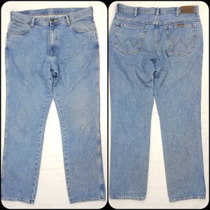May include: A pair of light blue denim jeans with a straight leg fit. The jeans have a classic five-pocket design and a brown leather patch on the back waistband with the Wrangler logo.