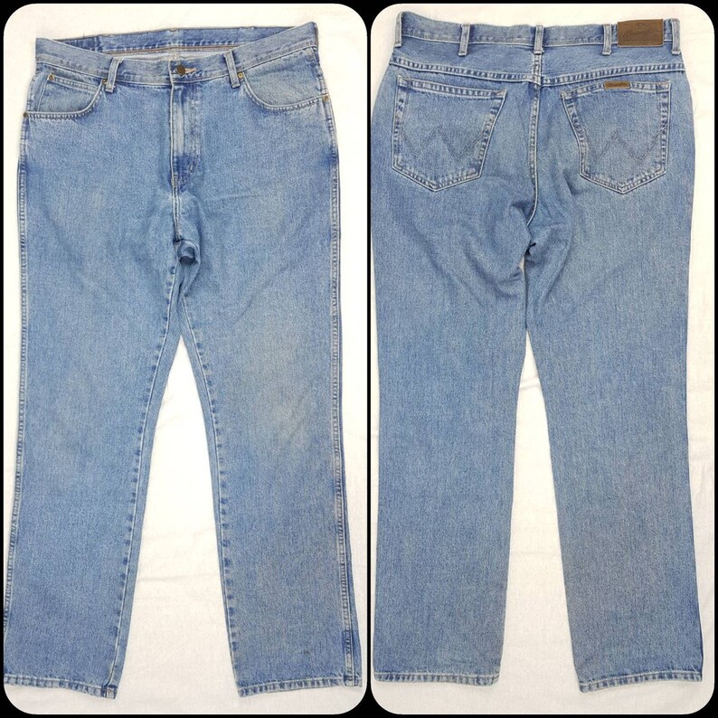 May include: A pair of light blue denim jeans with a straight leg fit. The jeans have a classic five-pocket design and a brown leather patch on the back waistband with the Wrangler logo.