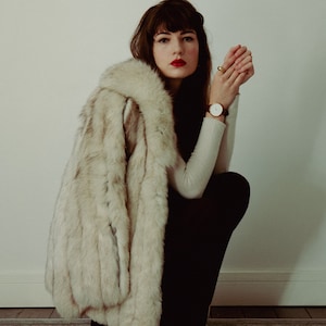 Op de afbeelding: Een vrouw draagt een witte nep bontjas met een zwarte turtleneck en een zwarte broek. Ze draagt een gouden horloge om haar linker pols en heeft haar handen voor haar gevouwen.