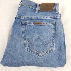 May include: A pair of blue denim jeans with a brown leather Wrangler label on the back pocket. The jeans are folded in half and have a light blue wash.