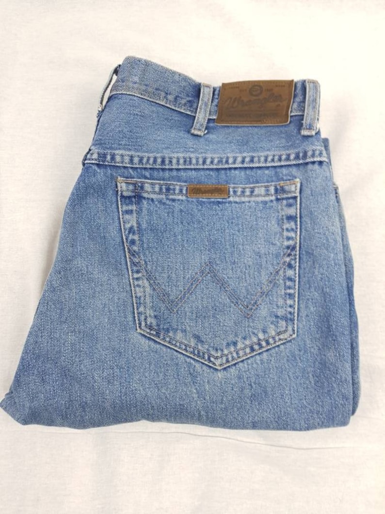 May include: A pair of blue denim jeans with a brown leather Wrangler label on the back pocket. The jeans are folded in half and have a light blue wash.