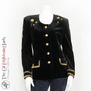 May include: A black velvet blazer with gold buttons and trim. The blazer has a round neckline and a single-breasted closure. The blazer is adorned with gold embellishments on the shoulders and pockets.
