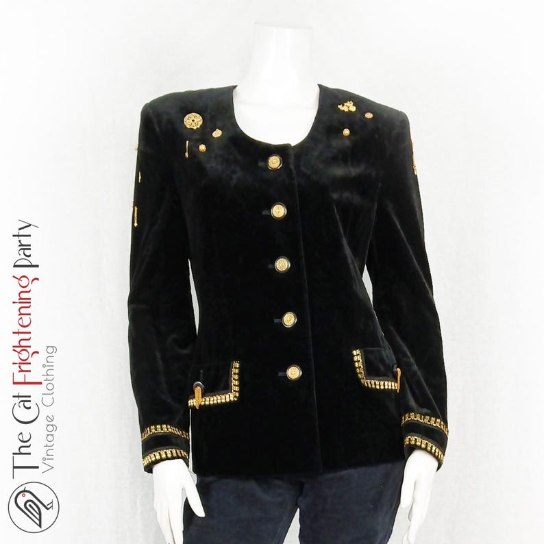 May include: A black velvet blazer with gold buttons and trim. The blazer has a round neckline and a single-breasted closure. The blazer is adorned with gold embellishments on the shoulders and pockets.