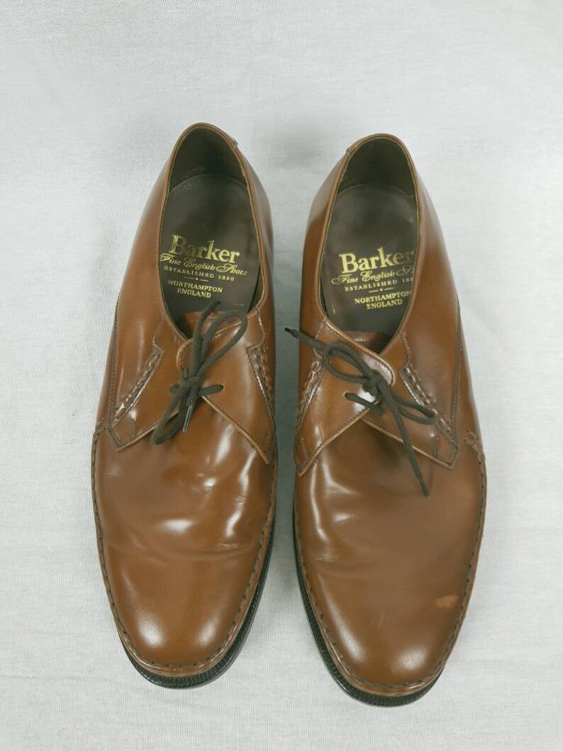 May include: A pair of brown leather lace-up shoes with a black sole. The shoes have a label on the inside that reads "Barker Fine English Shoes Established 1880 Northampton England".