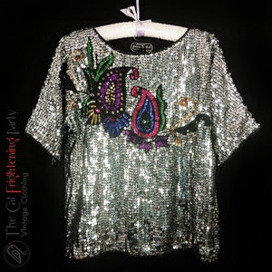 Può includere: Top argentato con paillettes e maniche corte, impreziosito da un colorato motivo floreale e paisley. Il top è appeso a una gruccia bianca su sfondo nero. Il logo The Cat Frightening Party Vintage Clothing è visibile.