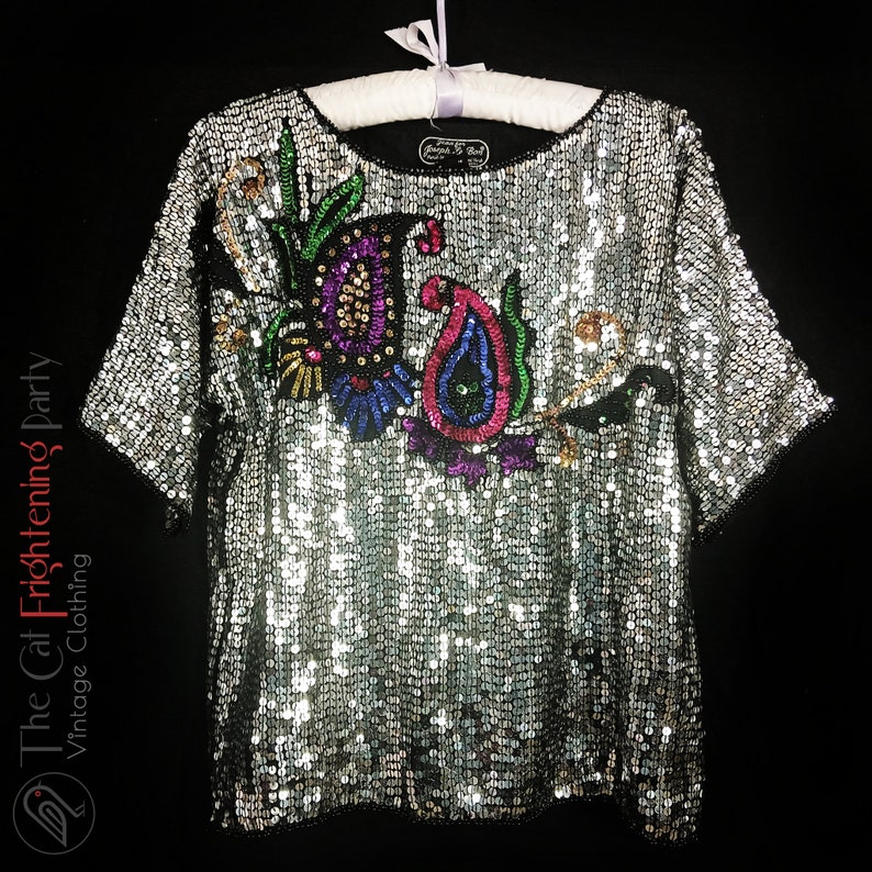 Pu&ograve; includere: Top argentato con paillettes e maniche corte, impreziosito da un colorato motivo floreale e paisley. Il top &egrave; appeso a una gruccia bianca su sfondo nero. Il logo The Cat Frightening Party Vintage Clothing &egrave; visibile.