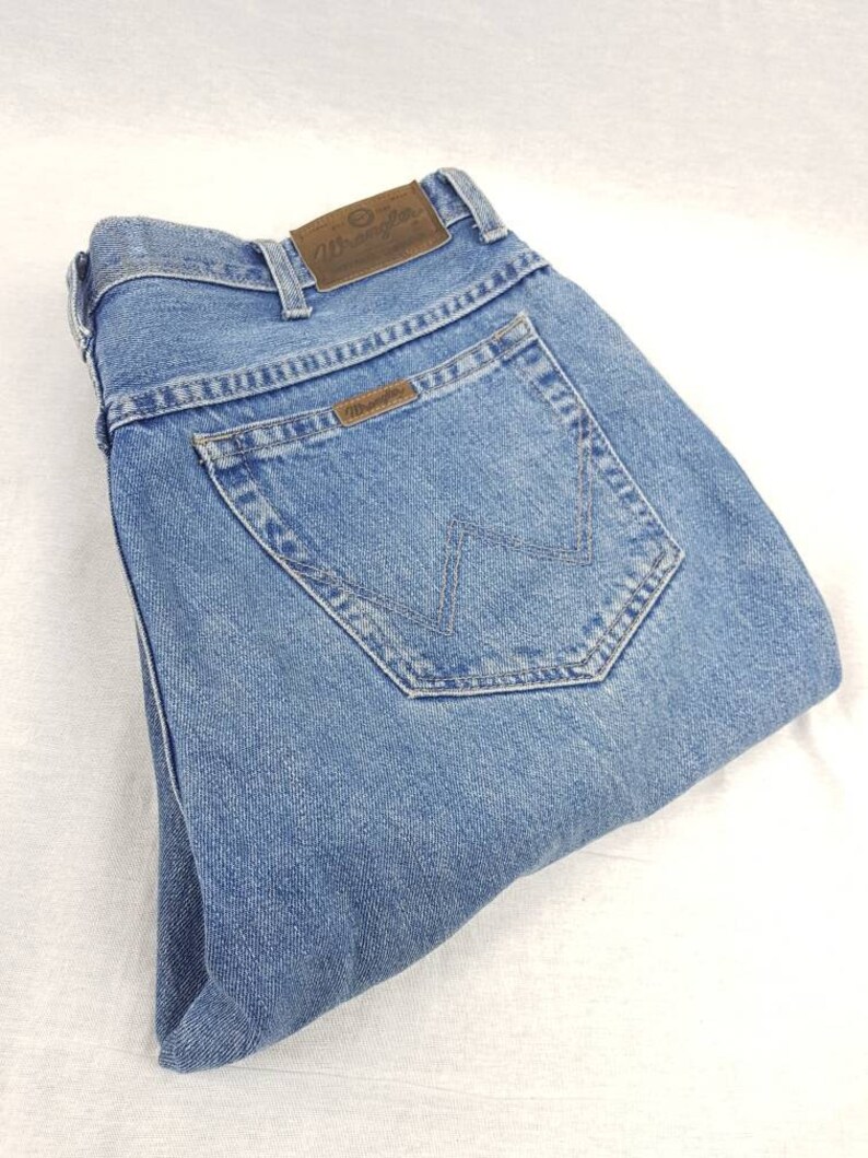 May include: A pair of light blue denim jeans with a brown leather Wrangler label on the back pocket.