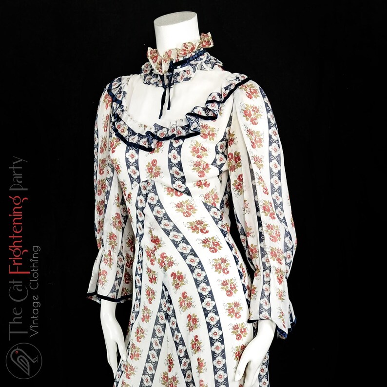 May include: A white vintage dress with a floral print and blue trim. The dress has a high neck and long sleeves with ruffles at the cuffs. The dress is made of a sheer fabric.